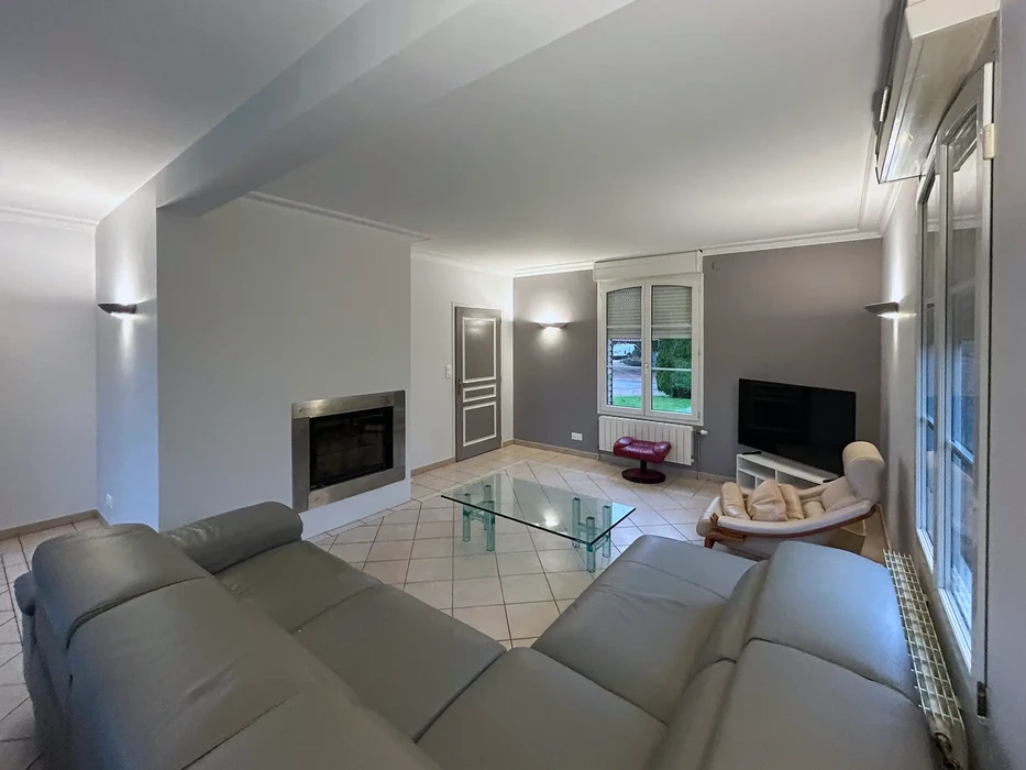 Salon moderne avec canapé d'angle gris, fauteuil beige, table basse en verre trempé sur piètement transparent