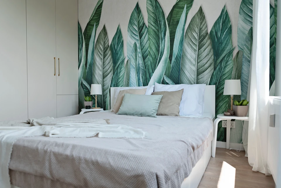 Chambre moderne avec lit double à tête de lit blanche, papier peint tropical aux motifs de feuilles vertes couvrant le mur principal